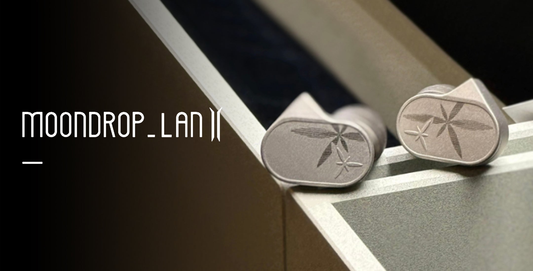 A simple, elegant shot of two MOONDROP LAN II earpieces resting on a beige-colored surface, possibly the interior packaging or case. The earpieces are in focus, showcasing their silver color and the laser-engraved designs (one appearing more star-like, the other more leaf-like).