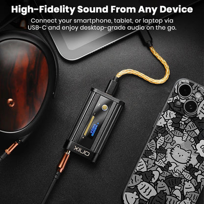 The black ONIX audio device connected via a gold-colored USB-C cable to a tablet or laptop, with the other end connected to a pair of large headphones, all resting on a dark surface next to a phone with a patterned case. 